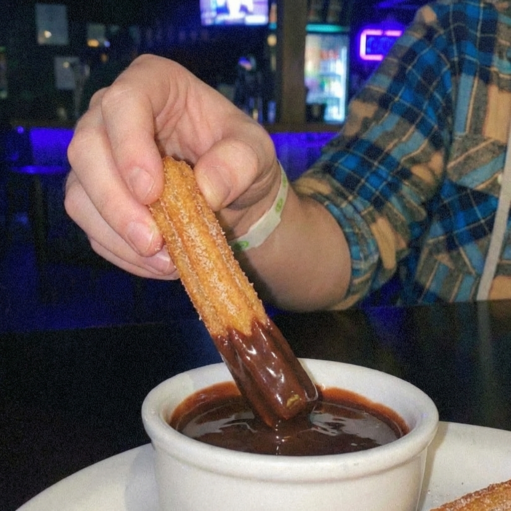 Close-up of a hand dipping a cinnamon sugar churro into glossy chocolate sauce
