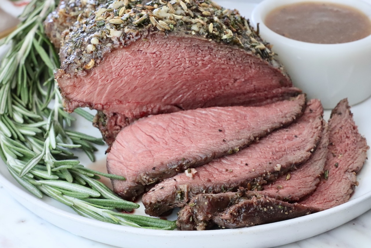 Close-up of a raw sirloin tip roast on a cutting board being rubbed with a garlic, rosemary, and Dijon mixture