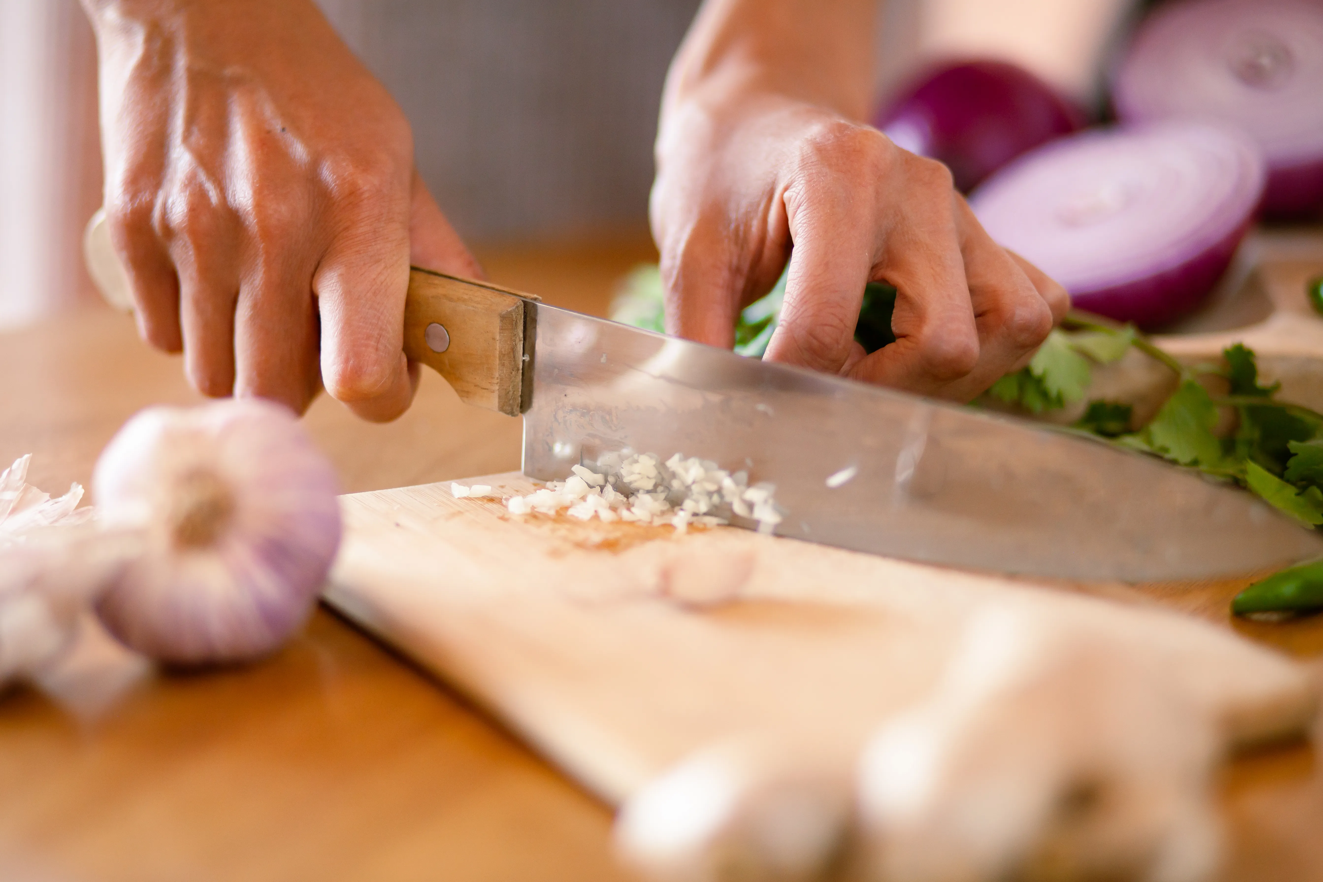 Close-up of hands slicing garlic cloves on a wooden cutting board with a chef’s knife