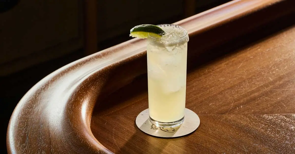 Club soda being poured into a tall glass cocktail over ice, with lemon garnish ready on the counter