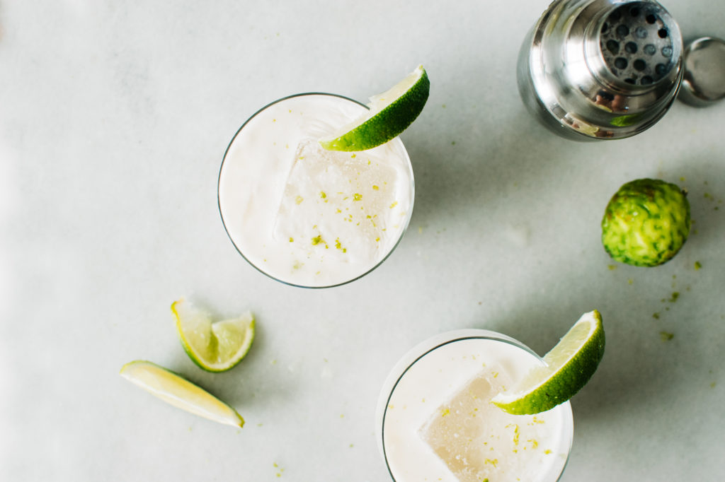 Coconut cream, tequila, limes, orange liqueur, agave, and a small bowl of salt arranged on a clean kitchen counter