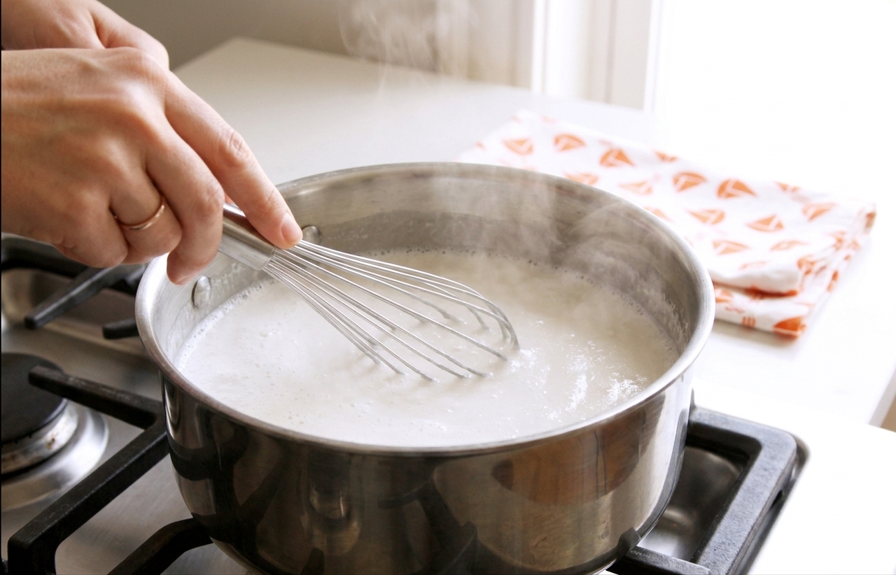 Coconut milk and sugar mixture steaming gently in a stainless steel saucepan on a stovetop while being whisked