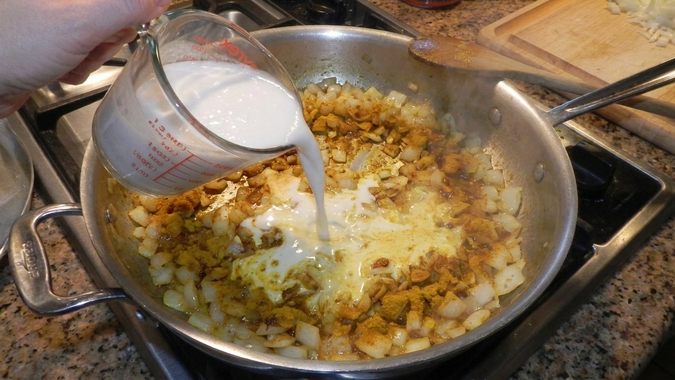 Coconut milk being poured into a skillet with onions and curry spices, creating a pale golden sauce