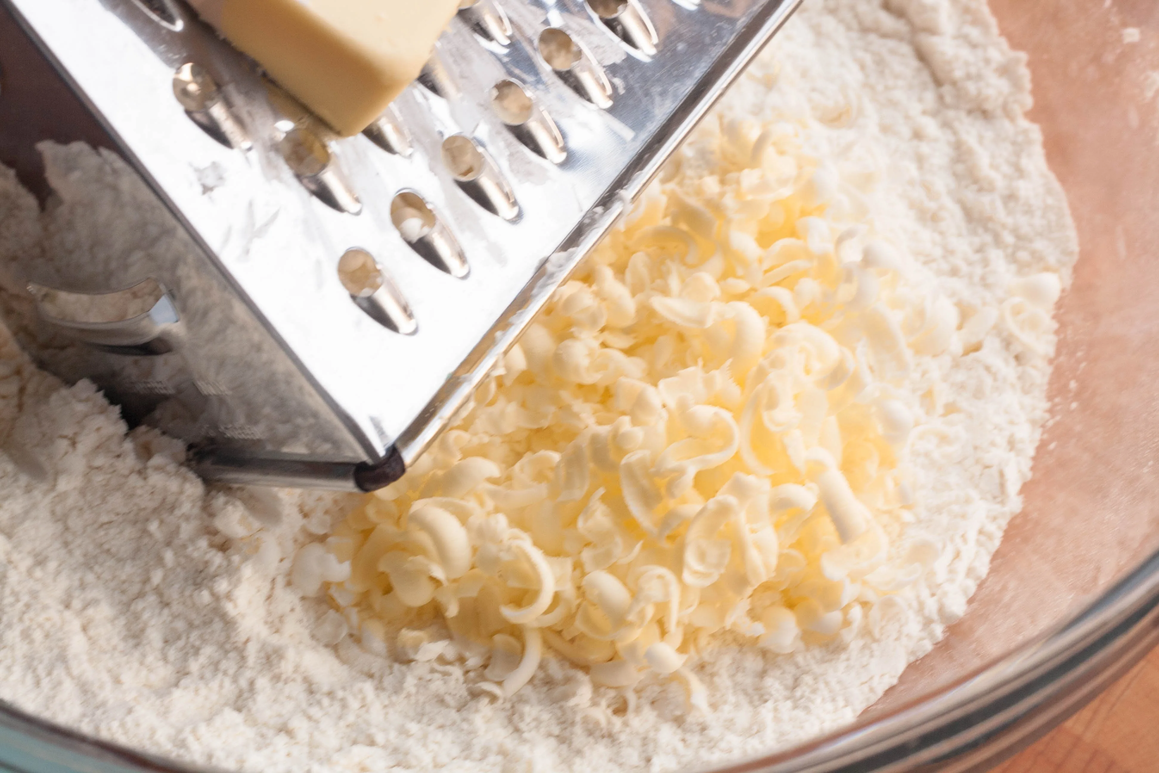 Cold butter being grated into a bowl of flour with a box grater on a kitchen counter