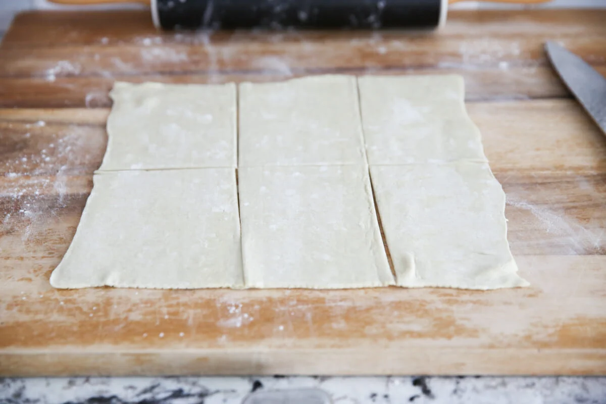 Cold puff pastry on a cutting board being cut into even squares with a chef's knife, photographed under soft kitchen lighting