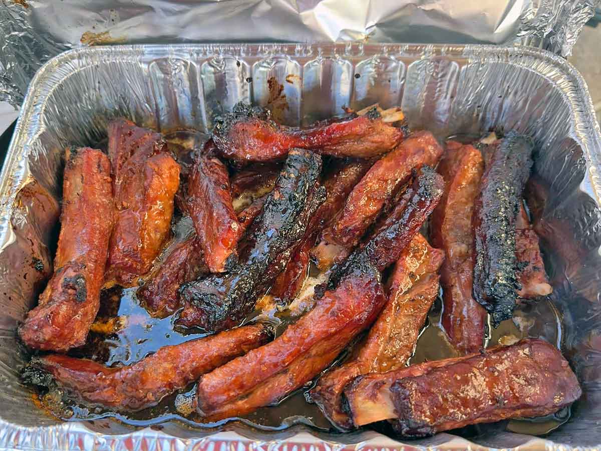Cooked BBQ riblets stored in a clear airtight container on a kitchen counter next to a small jar of extra barbecue sauce