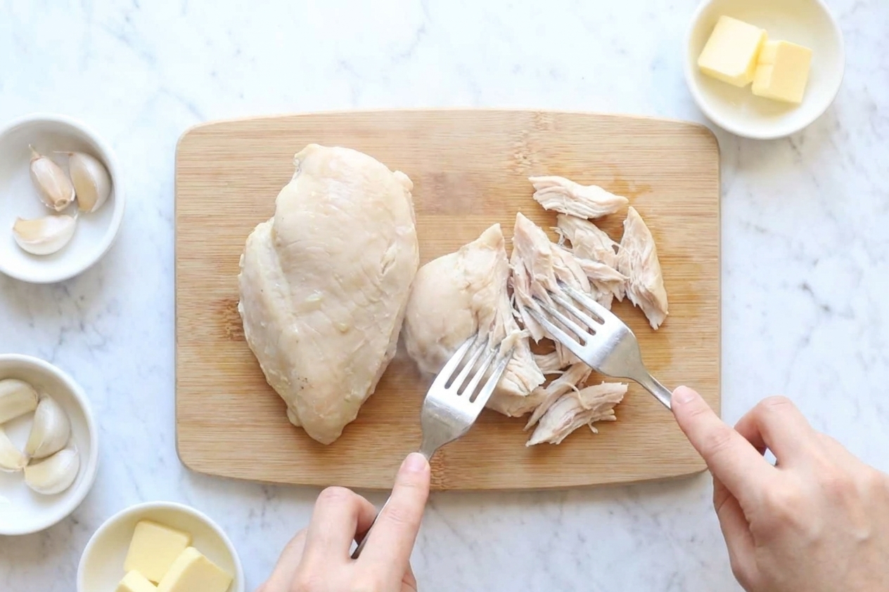 Cooked chicken being shredded with two forks on a cutting board