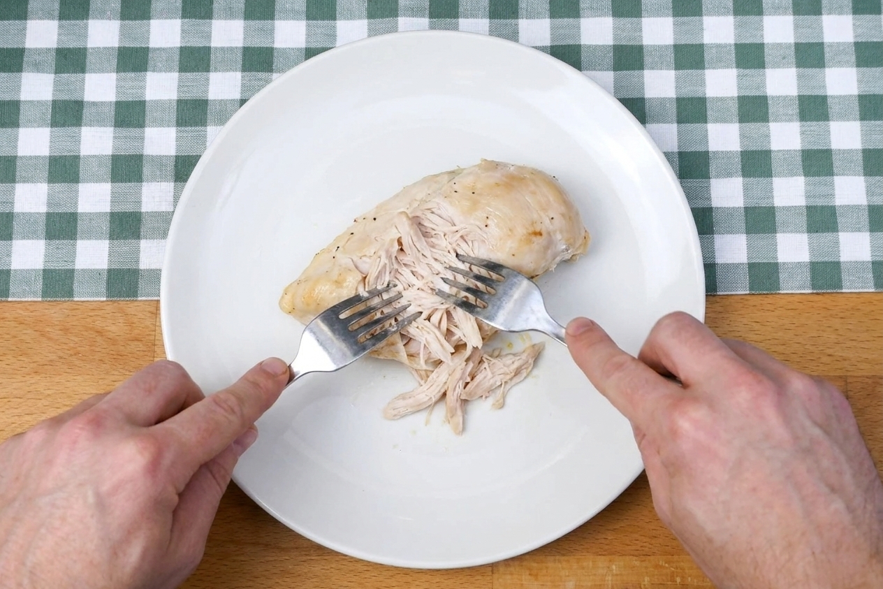 Cooked chicken being shredded with two forks on a white plate on a kitchen counter