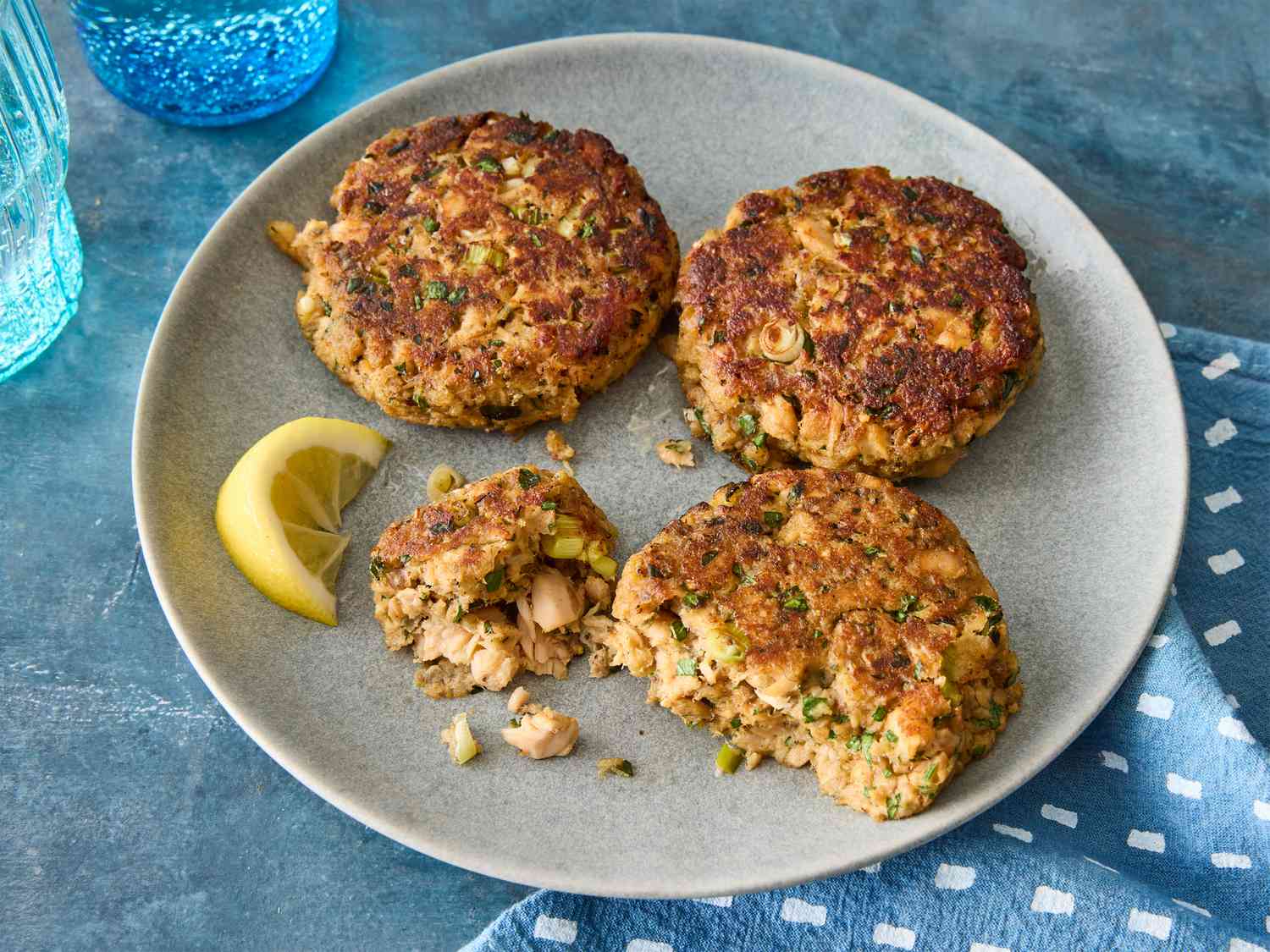 Cooked salmon patties stacked in an airtight glass container on a kitchen counter with a lemon half nearby