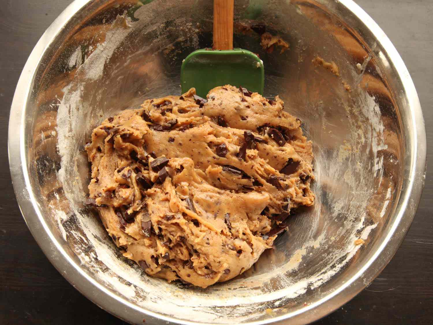 Cookie dough being folded with chocolate chips in a large mixing bowl using a rubber spatula on a home kitchen counter