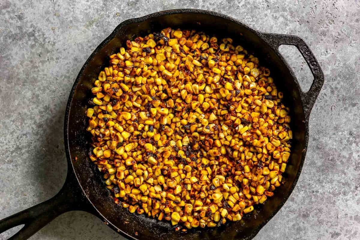 Corn kernels charring in a cast iron skillet on a stovetop with browned spots and a wooden spoon nearby