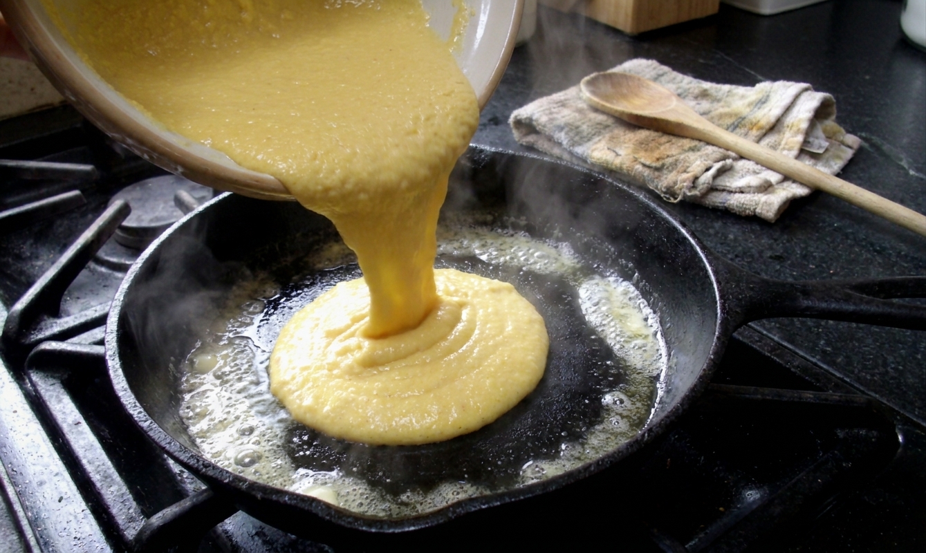Cornbread batter being poured into a buttered, hot cast iron skillet on a kitchen counter