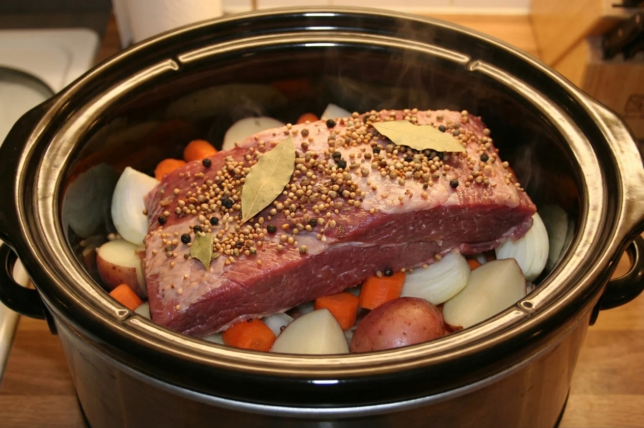 Corned beef brisket sitting in a slow cooker over onions, carrots, and potatoes with spices scattered on top