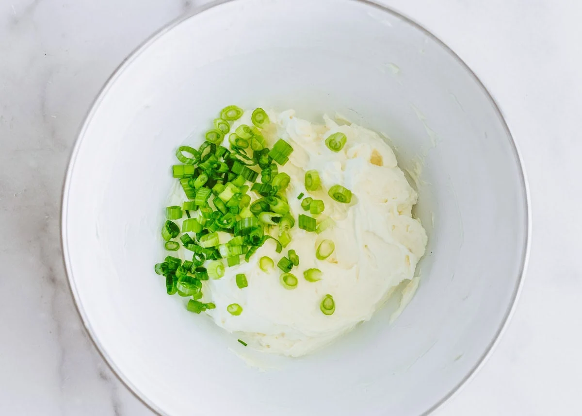 Cream cheese and sliced scallions in a mixing bowl with a spatula on a kitchen counter, ingredients nearby