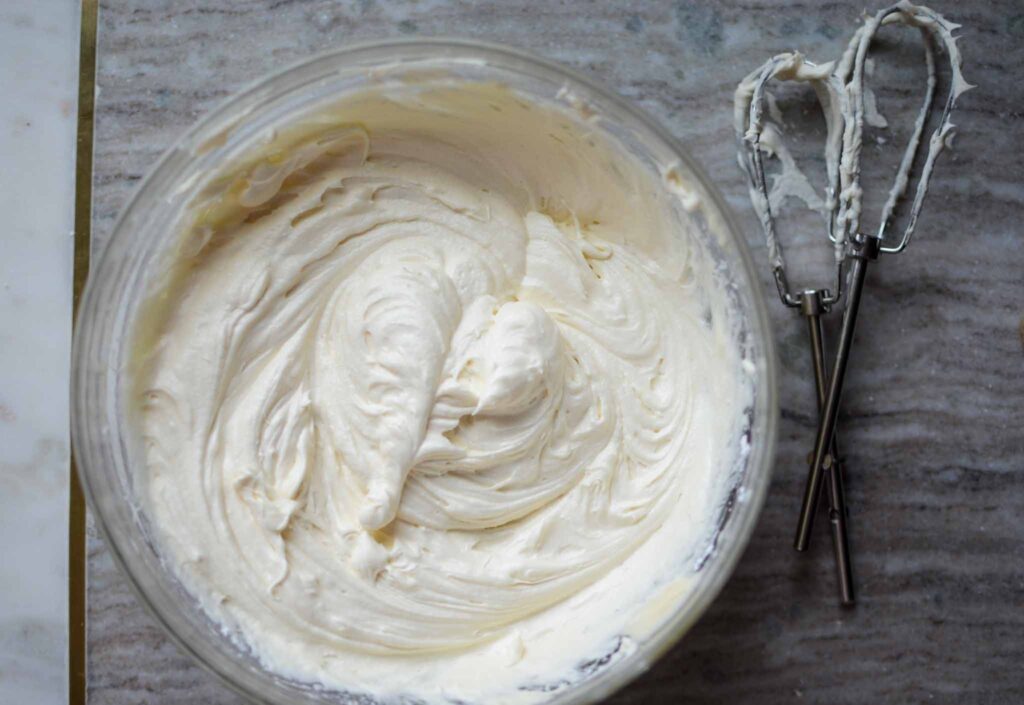 Cream cheese blocks, a bowl of granulated sugar, a small dish of vanilla extract, and lemons arranged on a clean kitchen counter.