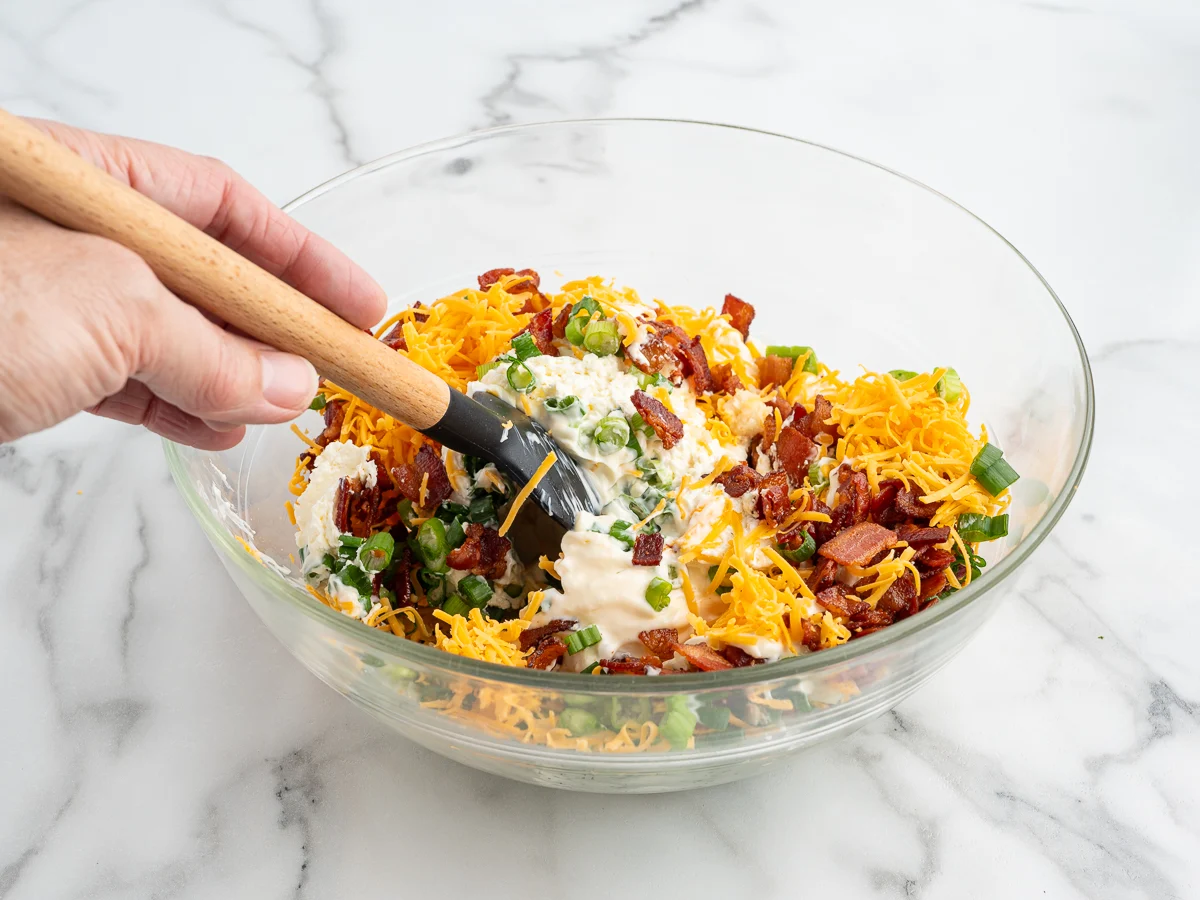 Crispy cooked bacon crumbles and freshly sliced green onions on a cutting board beside a bowl of shredded cheddar