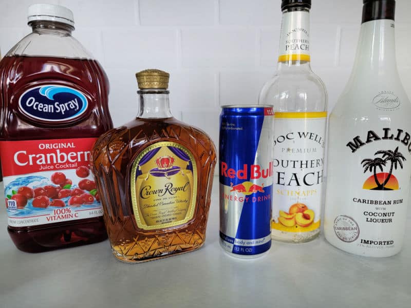 Crown Royal whisky, peach schnapps, Malibu coconut rum, and a small can of energy drink arranged on a clean kitchen counter with a jigger and cocktail shaker