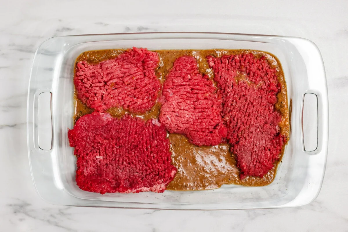 Cube steaks submerged in a vinegar and Worcestershire mixture inside a large glass bowl on a kitchen counter
