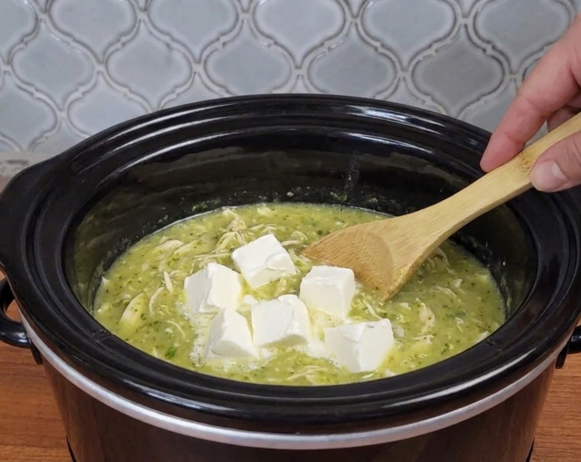 Cubed cream cheese softening in a slow cooker with salsa verde chicken, just before being stirred in