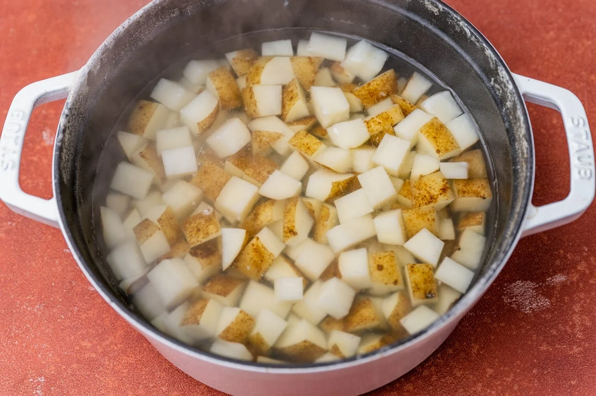 Cubed raw potatoes soaking in a clear glass bowl of water on a kitchen counter, with a towel and a knife nearby