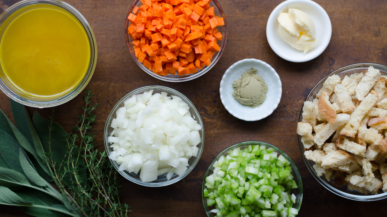 Cubes of dried bread, a bowl of chopped celery and onions, fresh herbs, eggs, and cartons of broth arranged on a wooden cutting board on a bright kitchen counter