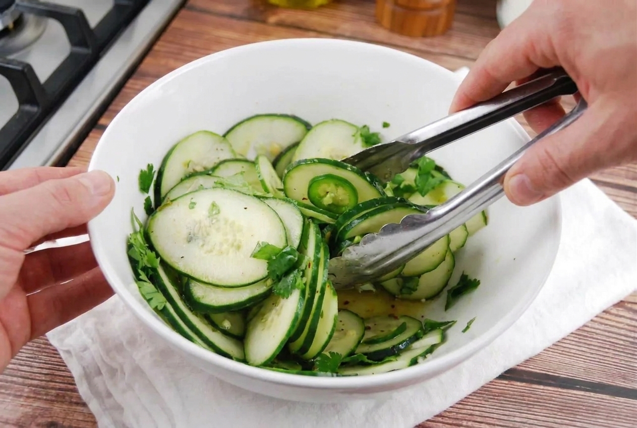 Cucumber salad being tossed with cilantro and sliced jalapeño in a large bowl using tongs on a kitchen counter