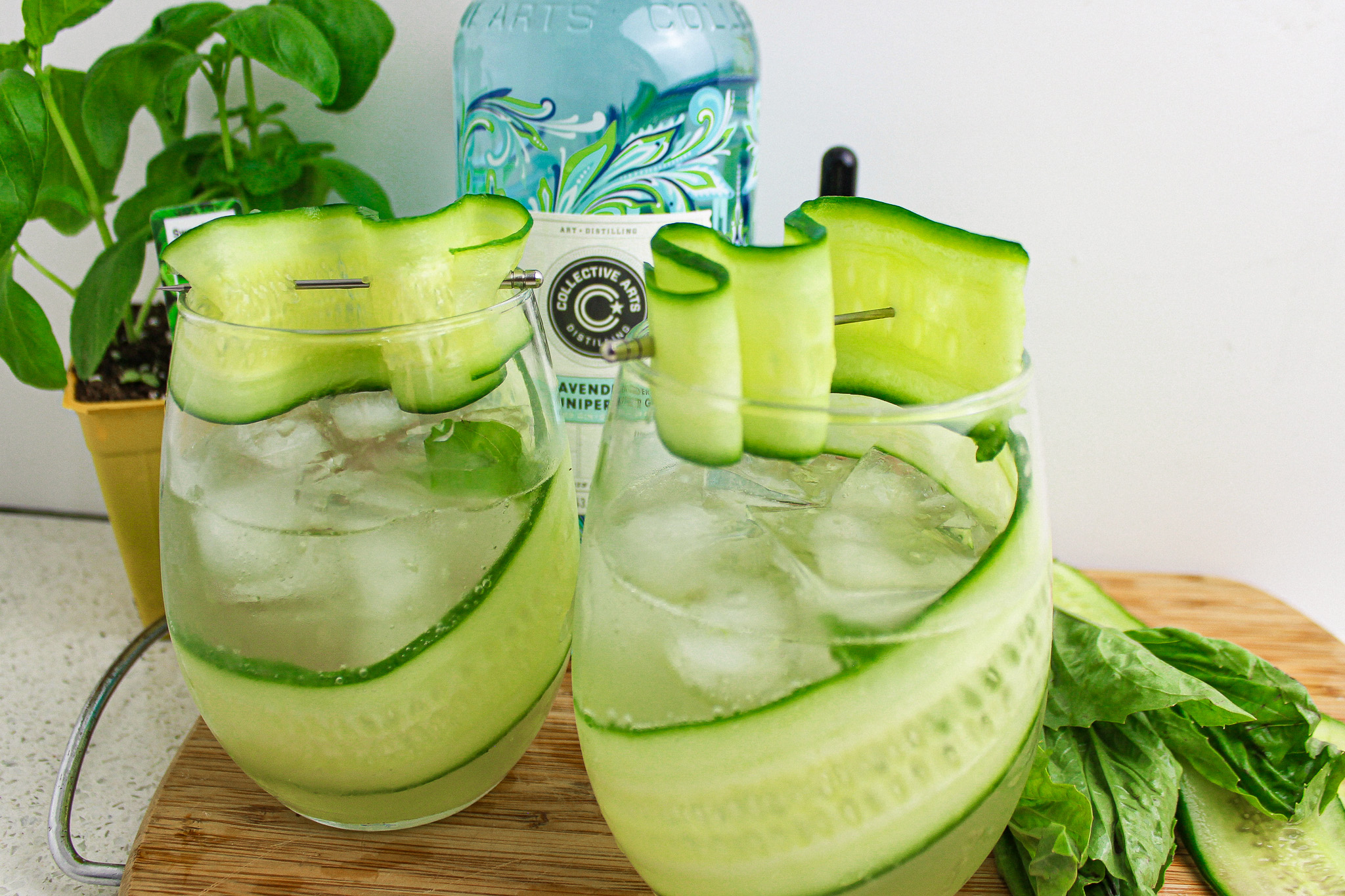 Cucumber slices and basil leaves being gently muddled in a metal cocktail shaker on a bright kitchen counter