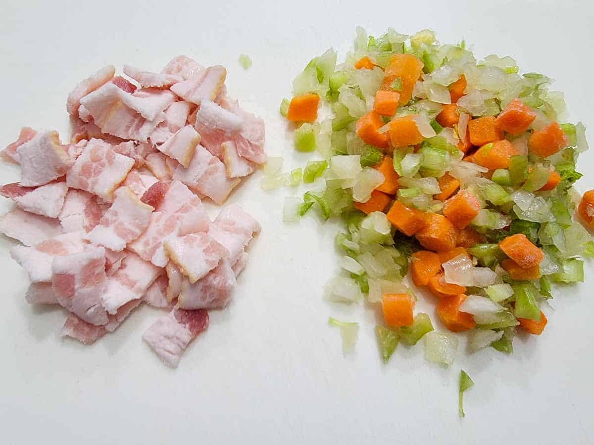 Diced onion and celery next to chopped bacon on a wooden cutting board on a home kitchen counter