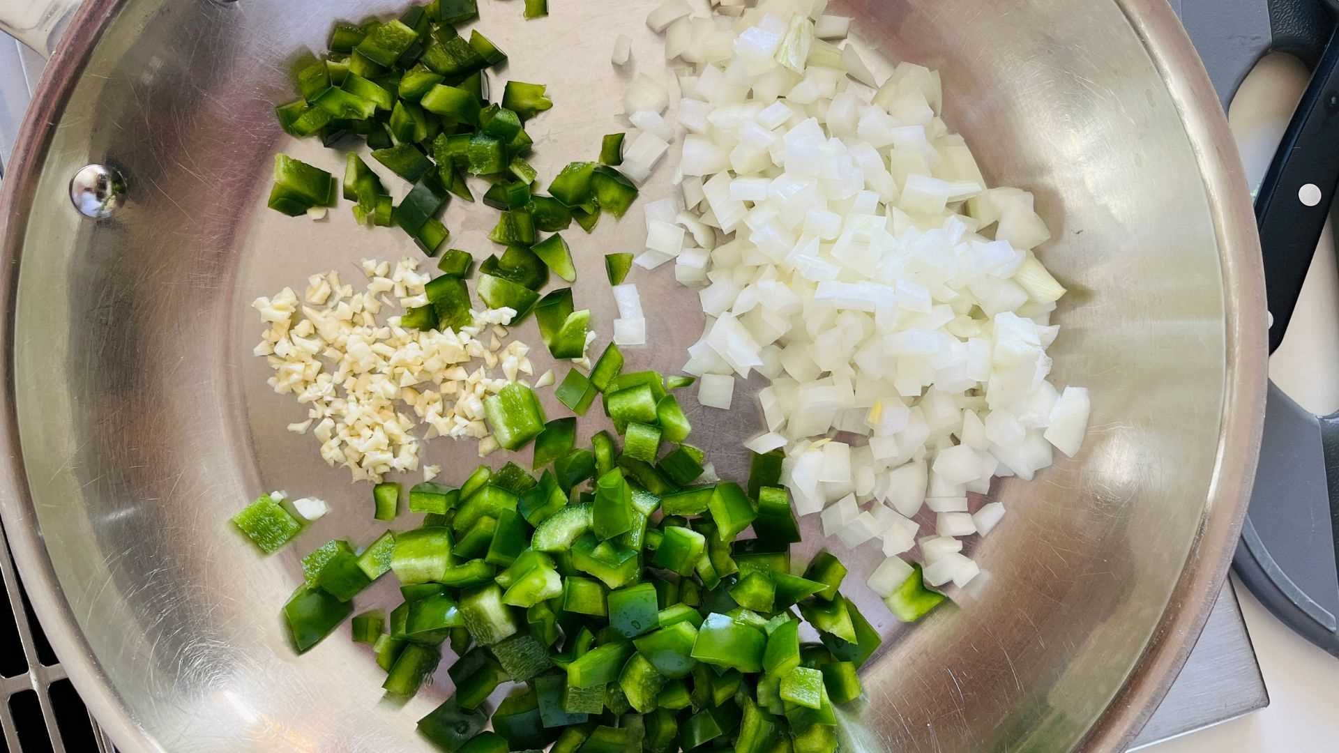 Diced onion and green bell pepper with minced garlic on a cutting board beside a skillet on a home kitchen counter