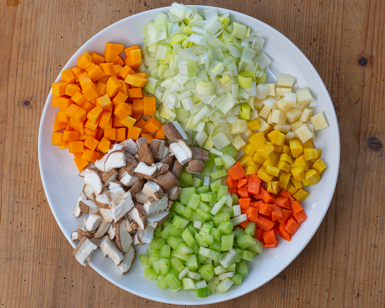Diced onion, carrots, celery, and potatoes on a wooden cutting board with a chef’s knife in a home kitchen