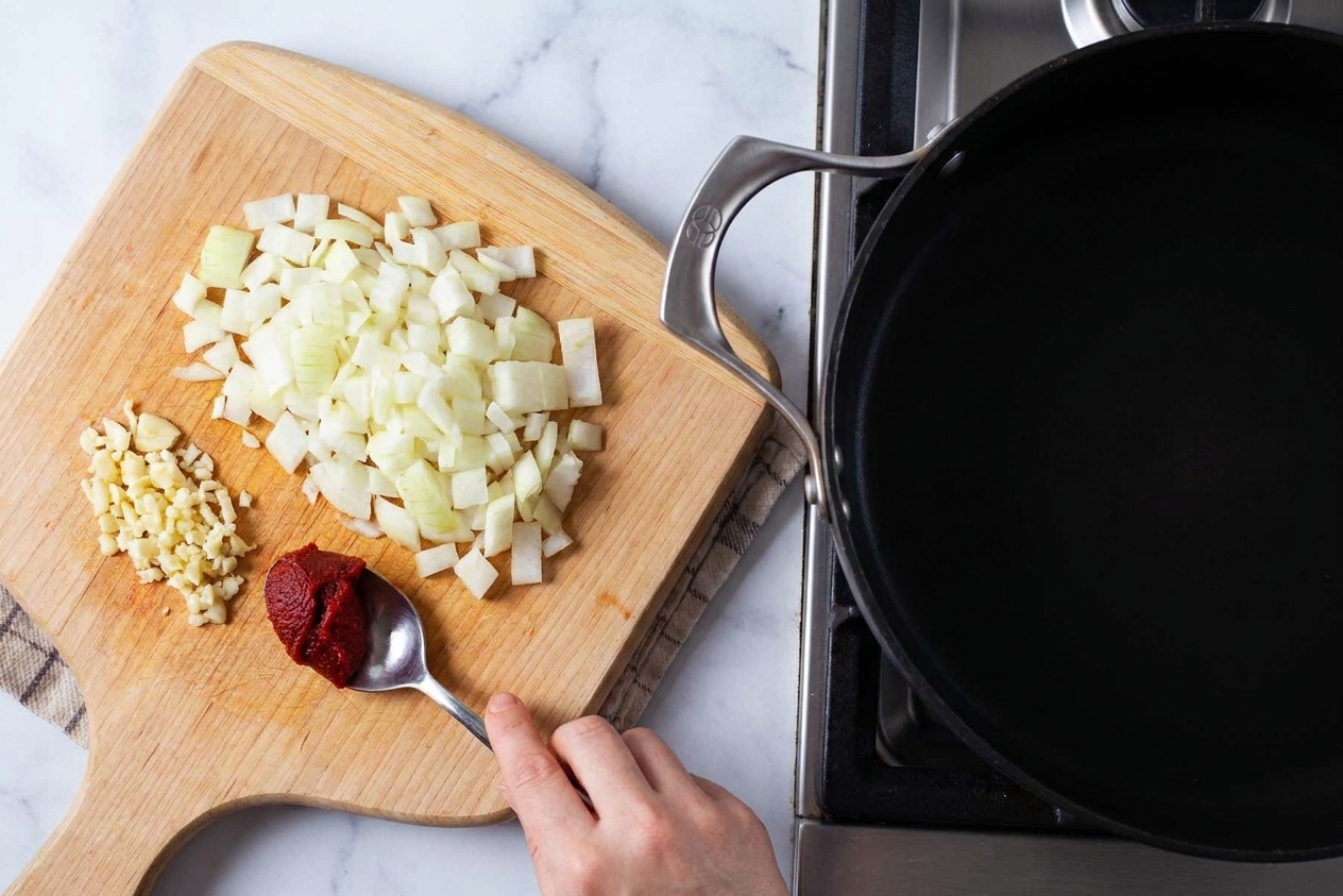 Diced onion, minced garlic, and a spoonful of tomato paste on a cutting board next to a skillet on a kitchen counter