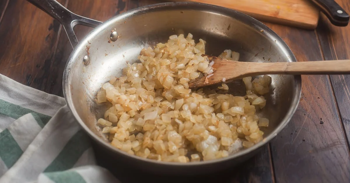 Diced onions sautéing in a large skillet on a stovetop with a wooden spoon