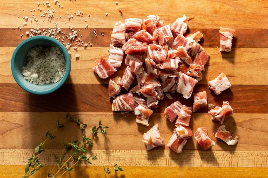 Diced pancetta on a wooden cutting board with a chef's knife and a small bowl of grated cheese nearby on a kitchen counter