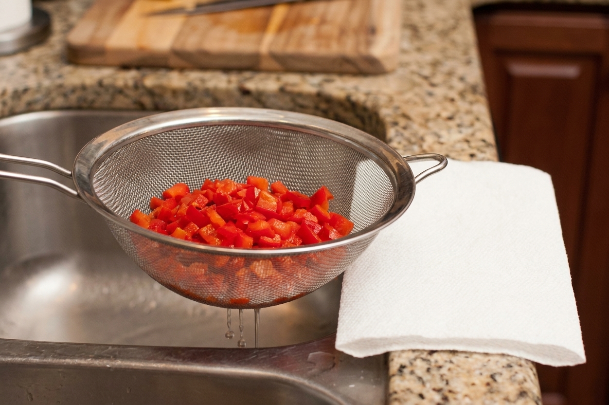Diced pimentos being drained in a fine mesh strainer over a sink with a paper towel nearby