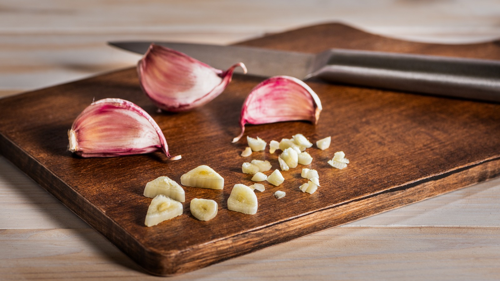 Diced yellow onion and minced garlic on a wooden cutting board beside a chef’s knife in a bright home kitchen