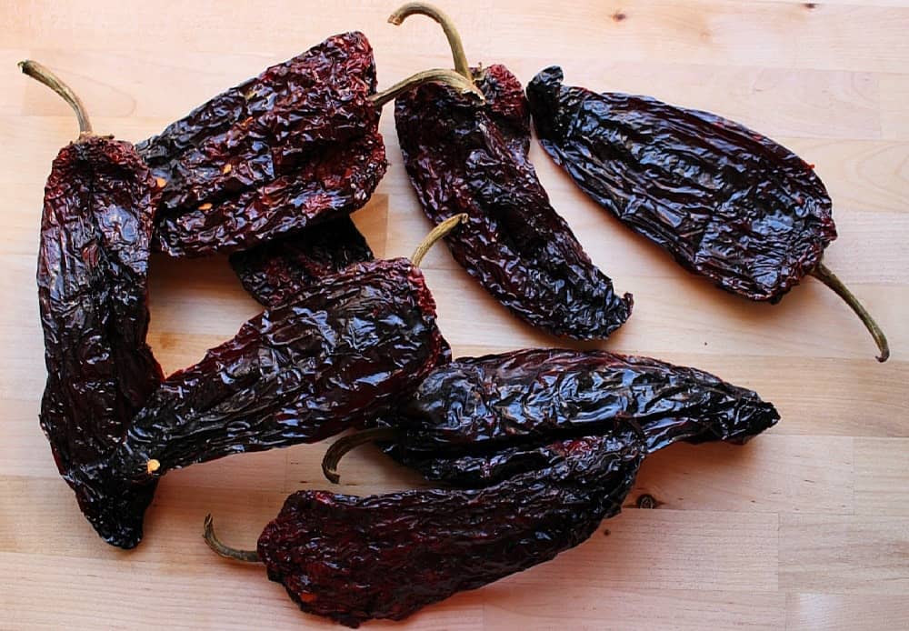 Dried ancho and pasilla chiles on a wooden cutting board beside peeled garlic and sliced onion on a kitchen counter