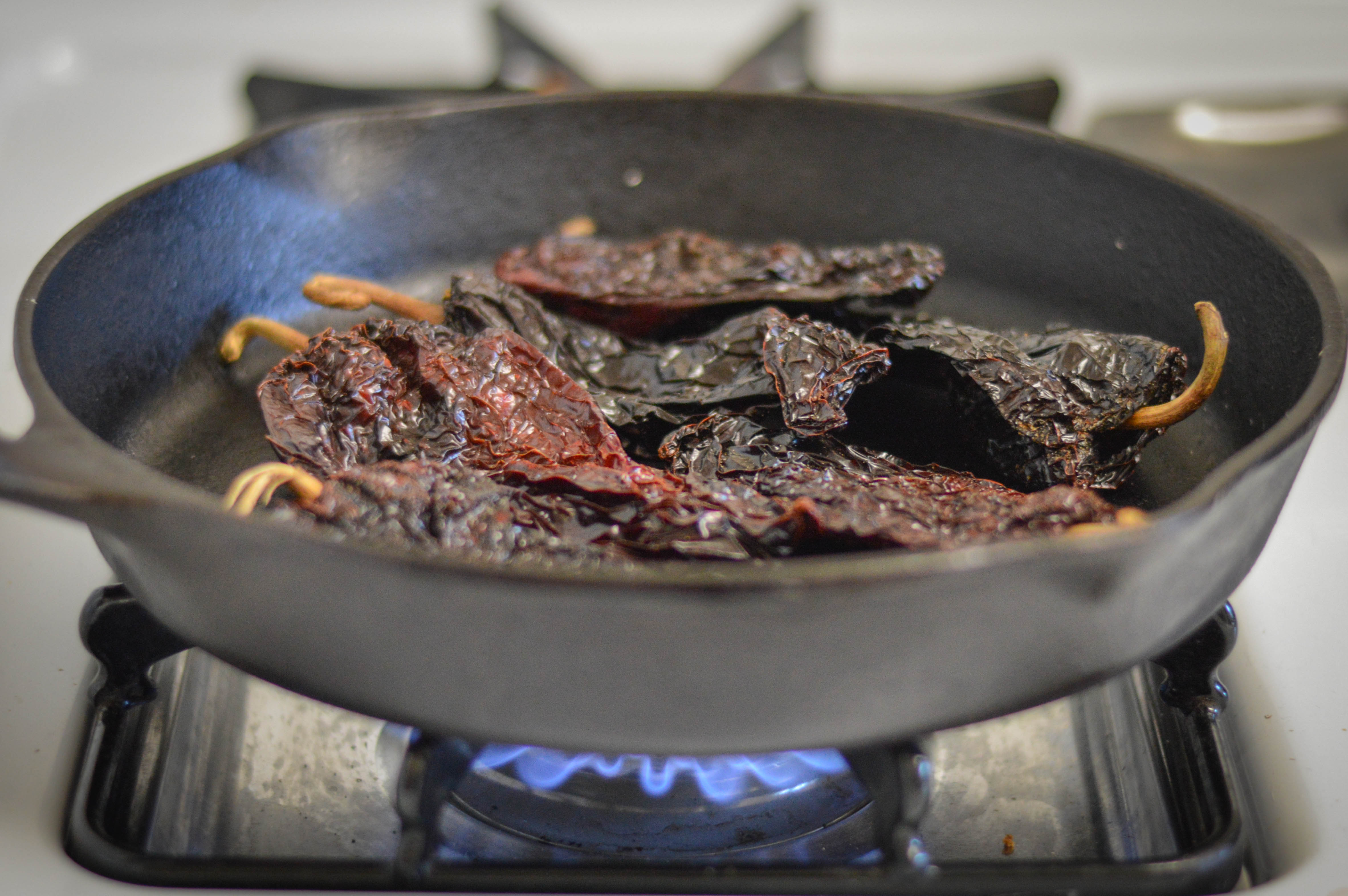 Dried ancho chiles toasting in a dry cast iron skillet on a home stove with tongs nearby