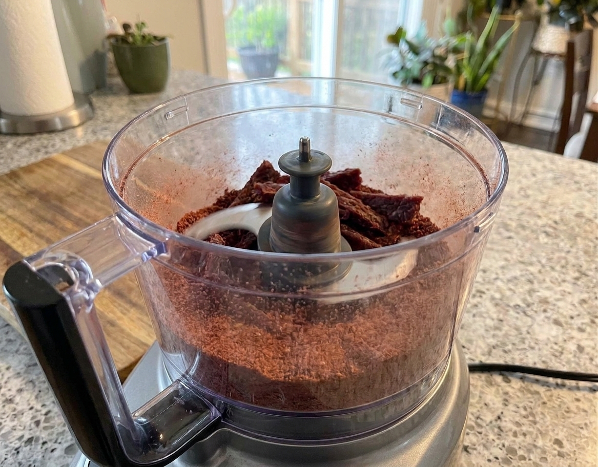 Dried beef being pulsed into a fine powder inside a food processor bowl on a kitchen counter