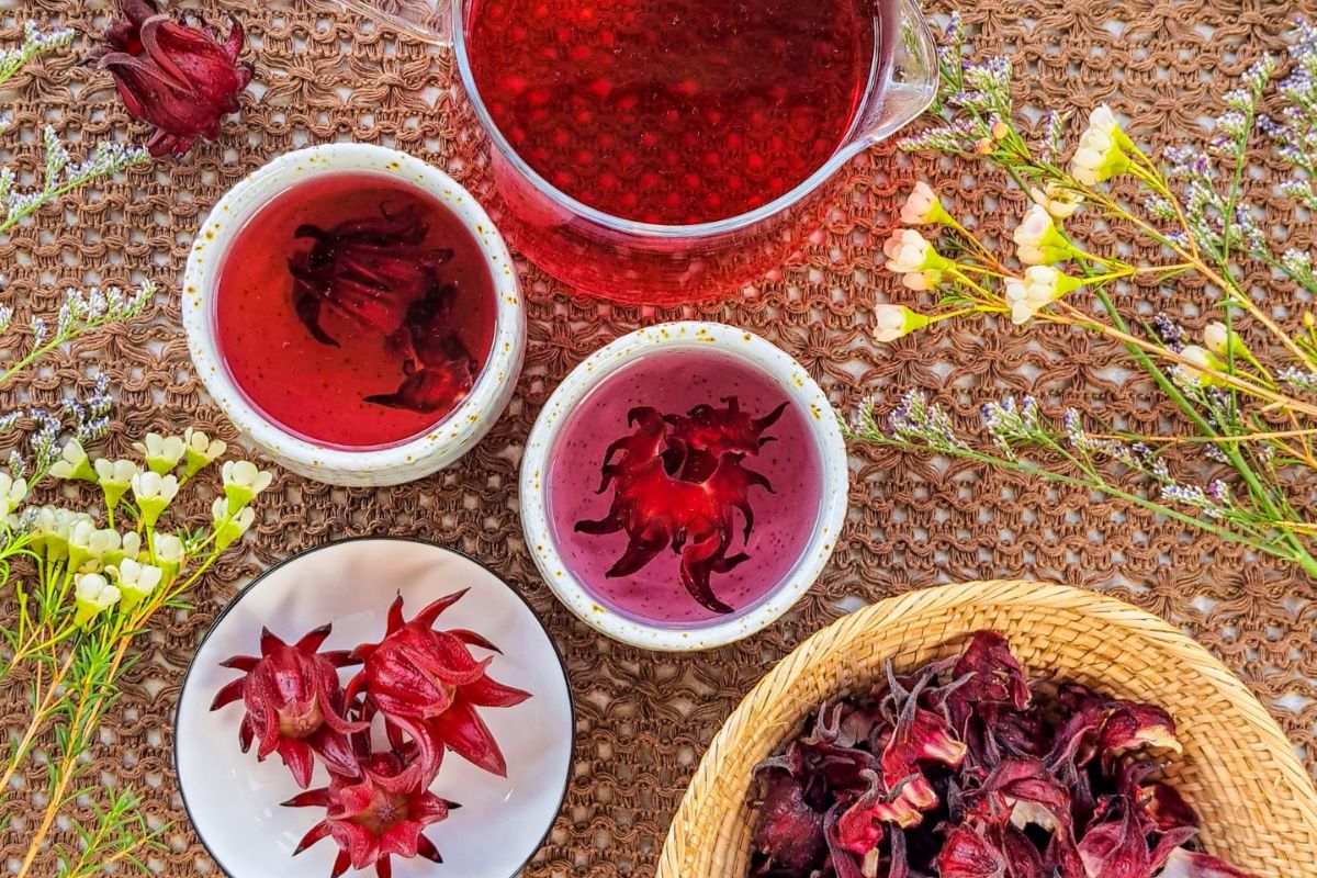 Dried hibiscus flowers, a cinnamon stick, limes, an orange, and a jar of honey arranged on a clean kitchen counter with measuring cups
