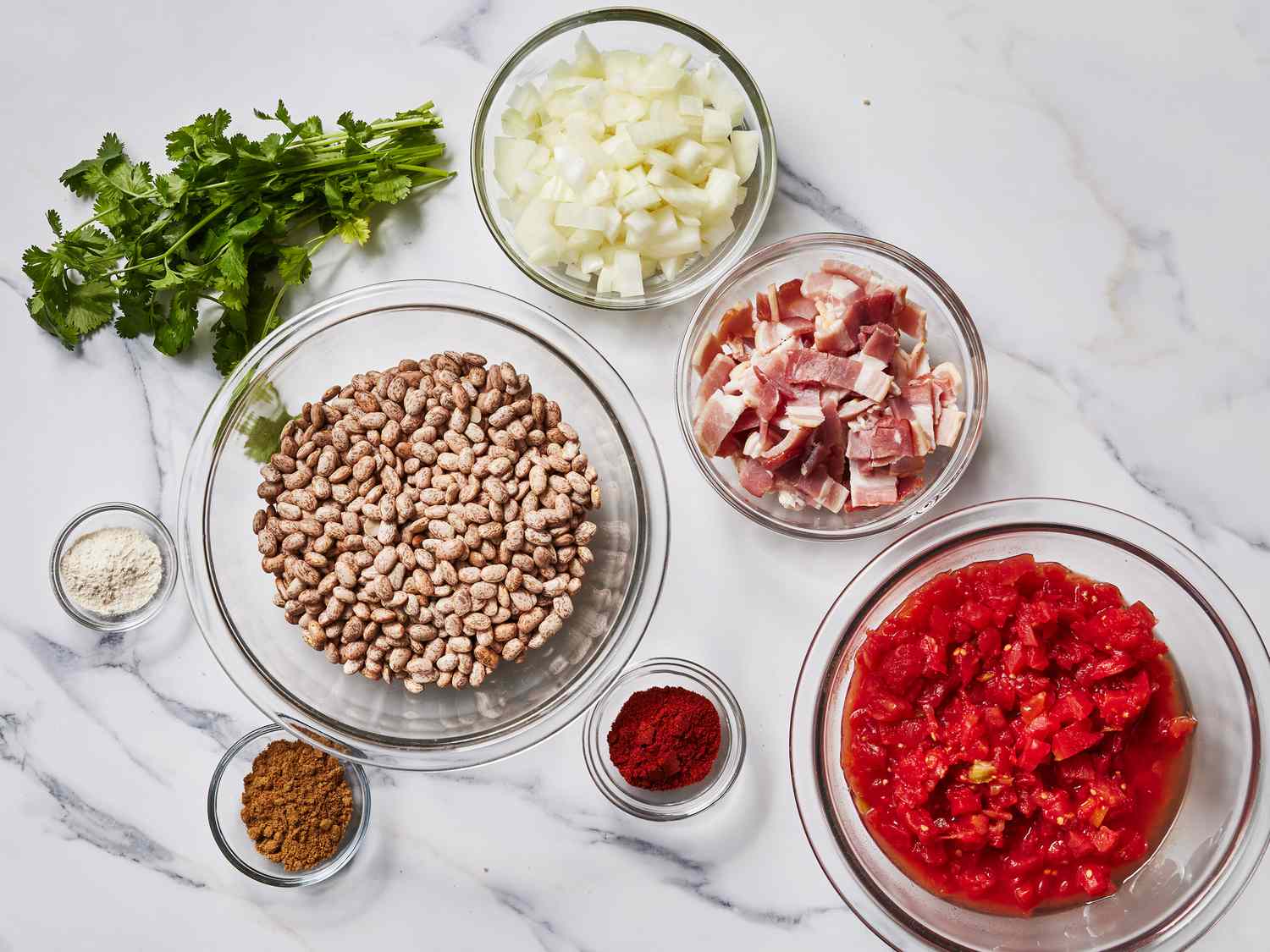 Dry pinto beans, onion, garlic, cumin, oregano, bay leaves, broth, and a small bowl of salt arranged on a clean kitchen counter in natural light