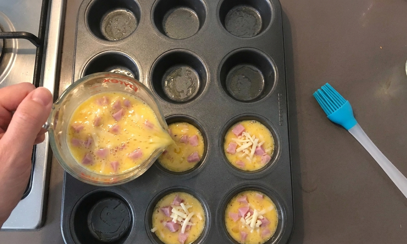Egg mixture being poured from a measuring cup into a greased muffin tin, with visible bits of cheese and ham