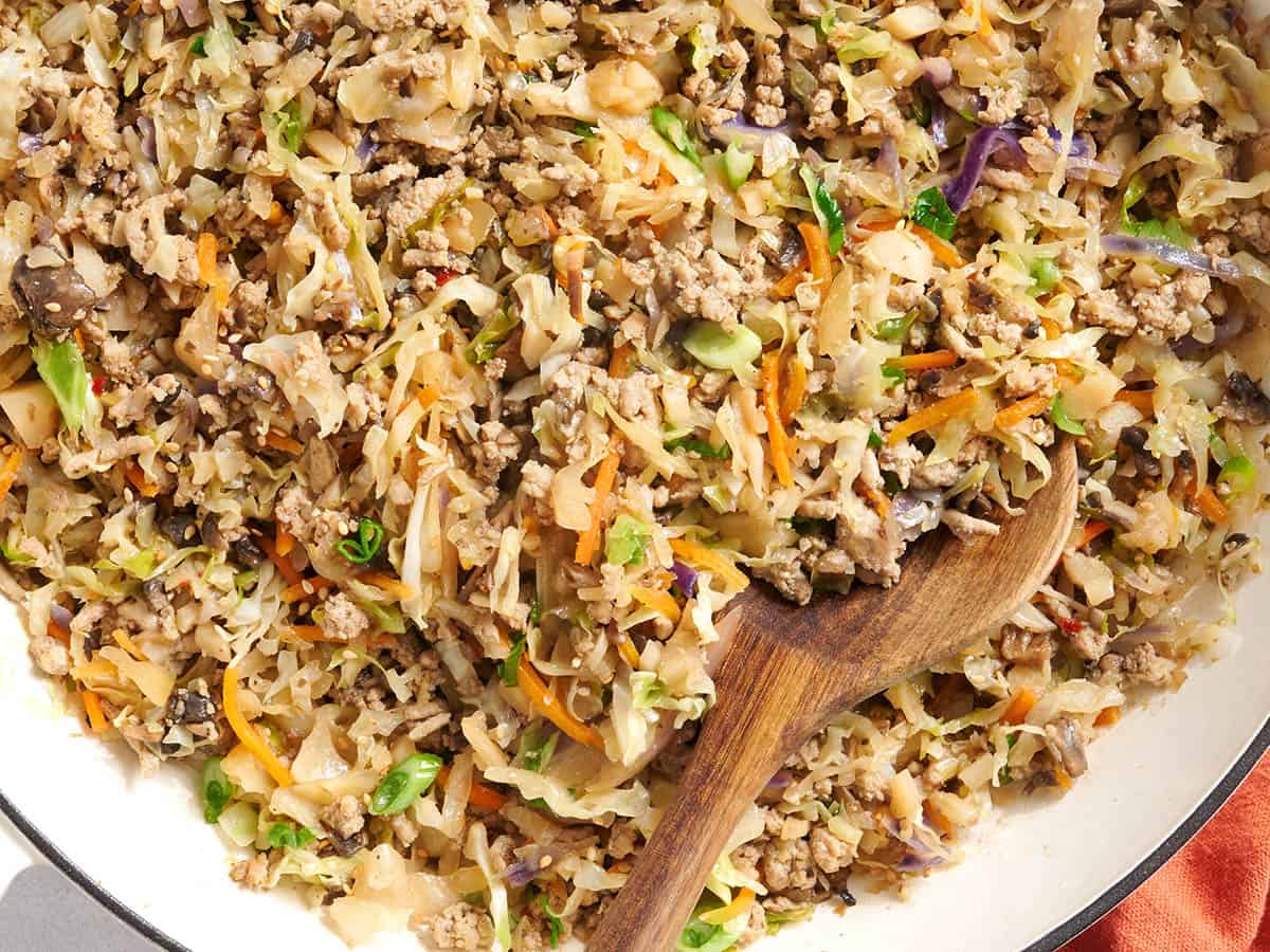 Egg roll in a bowl being stirred in a large skillet on a stovetop, with steam rising and a wooden spoon folding cabbage into browned meat