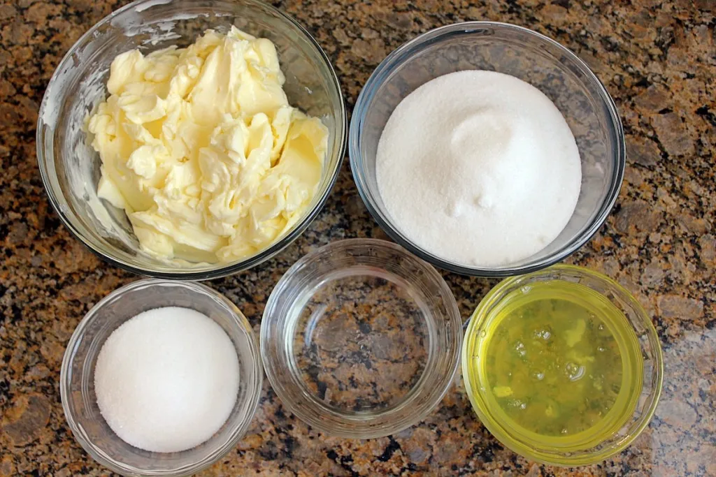 Egg whites in a small bowl next to granulated sugar, cubed butter, and vanilla on a clean kitchen counter