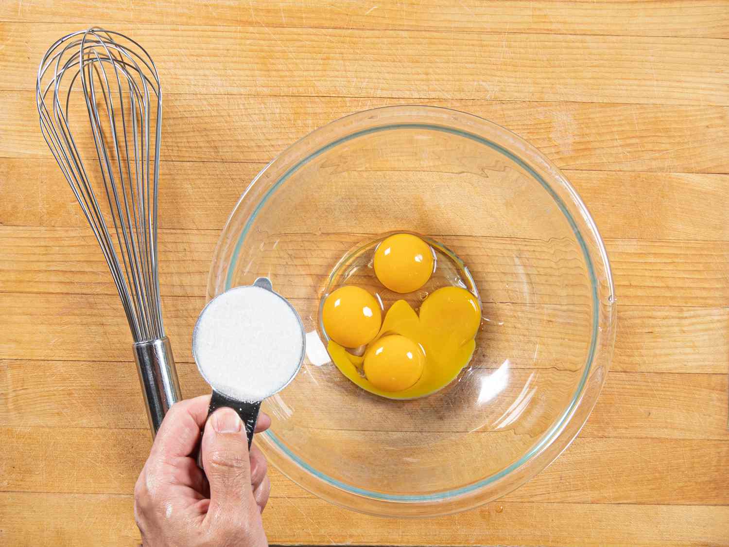 Egg yolks and sugar being whisked in a glass bowl on a kitchen counter with a whisk and small bowls nearby