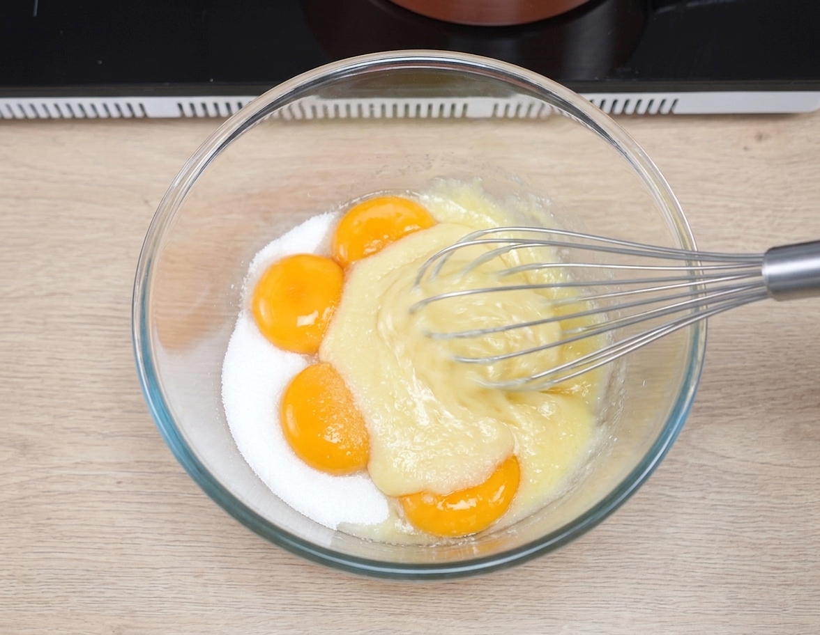 Egg yolks and sugar being whisked together in a glass bowl on a kitchen counter