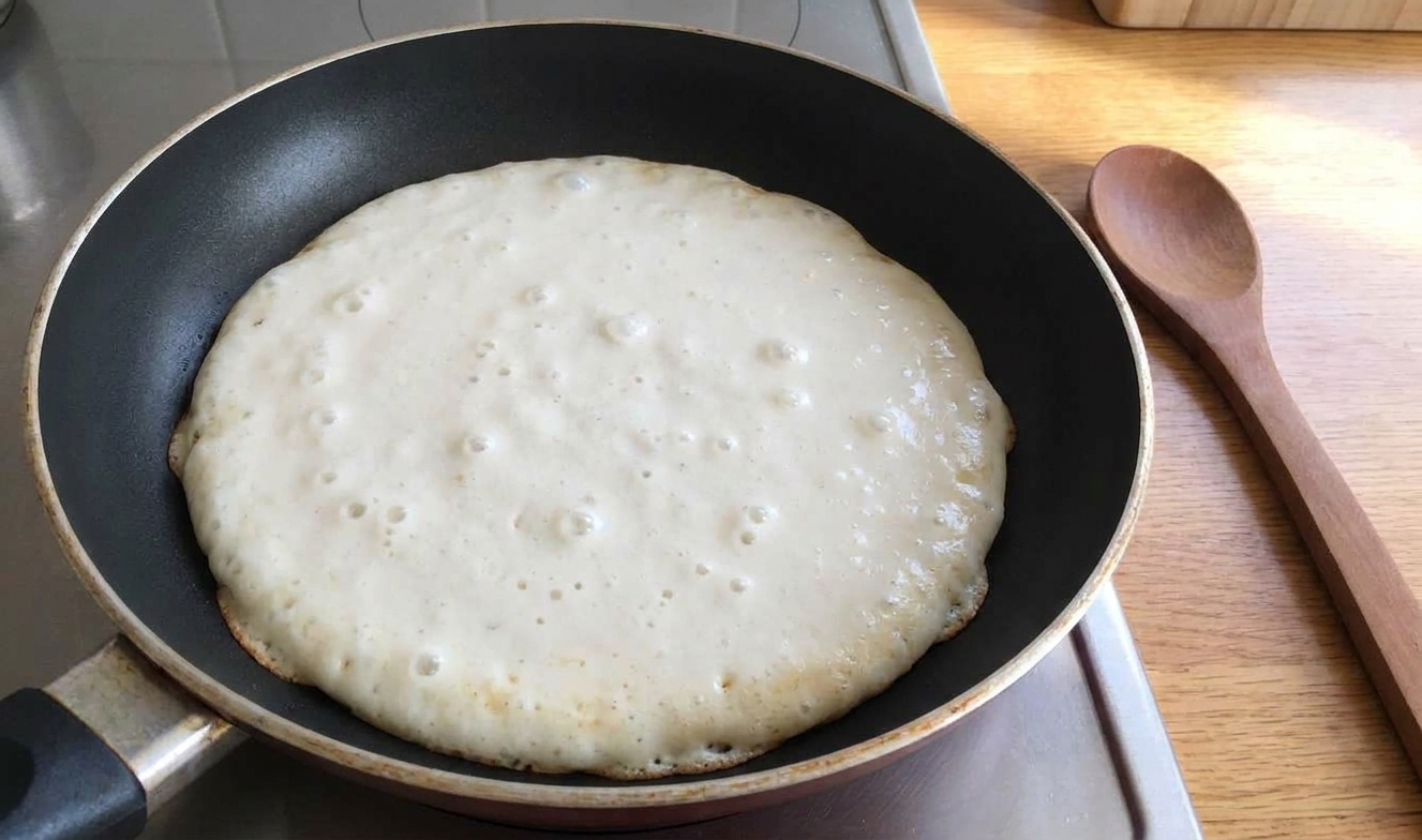 Eggless pancake batter cooking with small bubbles forming on the surface and the edges beginning to set
