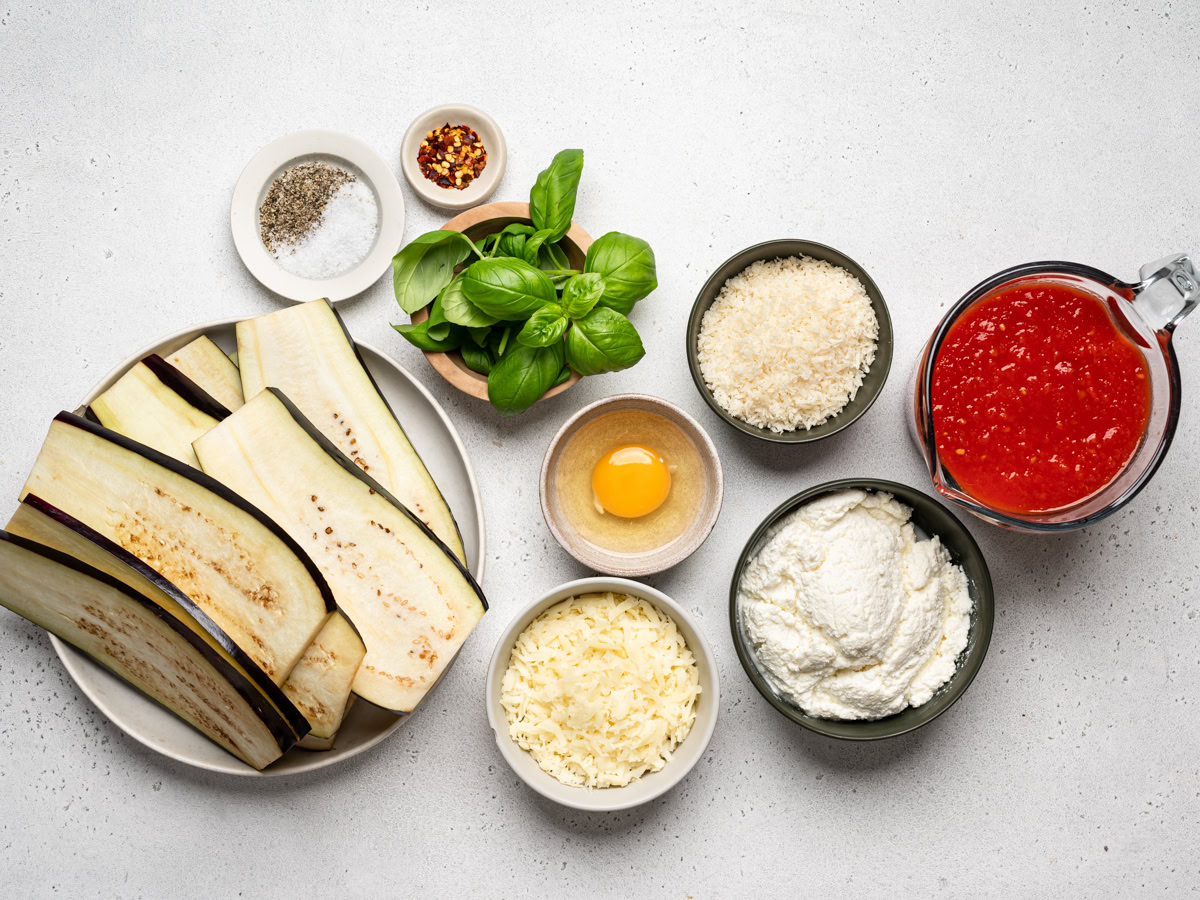 Eggplant, ricotta, mozzarella, Parmesan, eggs, garlic, basil, and a jar of marinara arranged on a clean kitchen counter with warm natural light