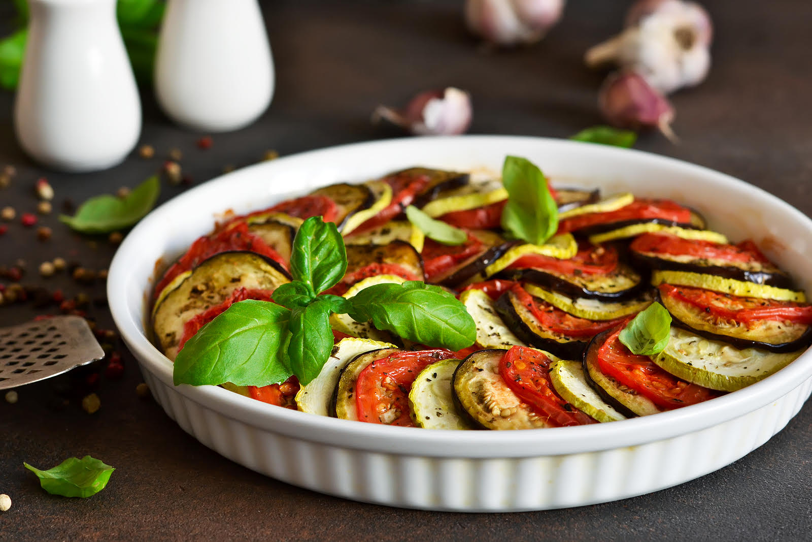 Eggplant, zucchini, yellow squash, tomatoes, bell peppers, garlic, onion, olive oil, and fresh herbs arranged together on a bright kitchen counter