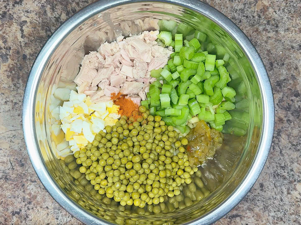 Elbow macaroni, canned tuna, mayonnaise, celery, red onion, pickles, mustard, and lemon arranged on a clean kitchen counter in consistent warm lighting