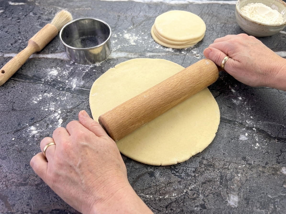 Empanada dough being rolled on a lightly floured counter with a round cutter nearby